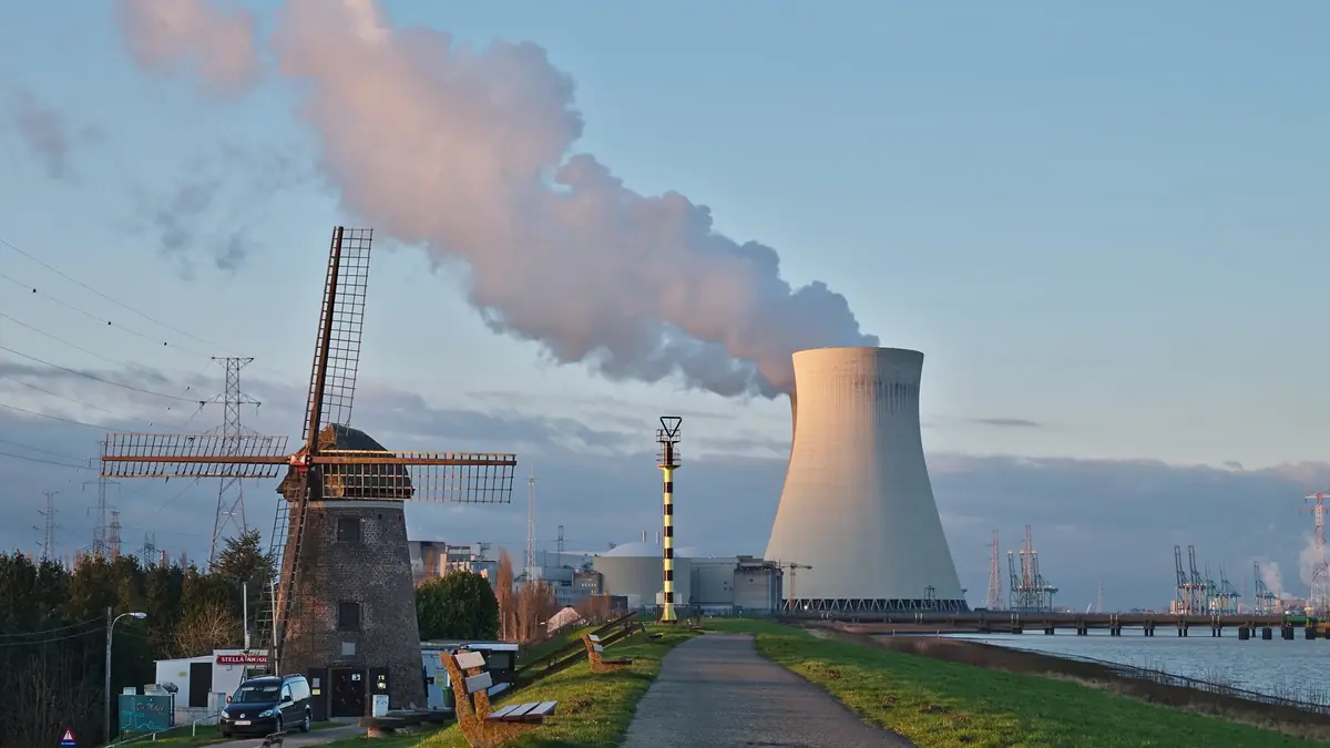 A nuclear power plant cooling tower venting steam at dusk beside a brick windmill and high-voltage pylons at Doel, Belgium